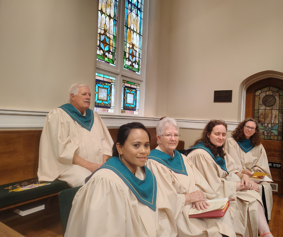 Chancel Choir members in cream robes with teal stoles seated in the sanctuary pews in front of a stained glass window