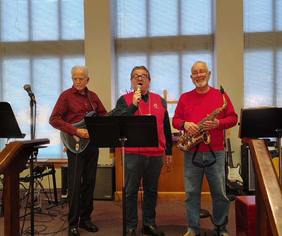 Three Praise Band musicians in red sweaters playing guitar, singing at a microphone and playing saxophone