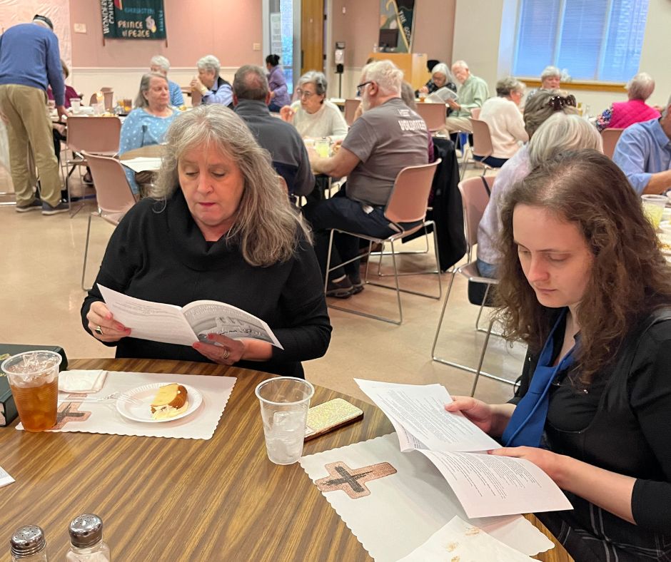 Two women reading documents at fellowship meal in church hall with congregation members dining at tables in background
