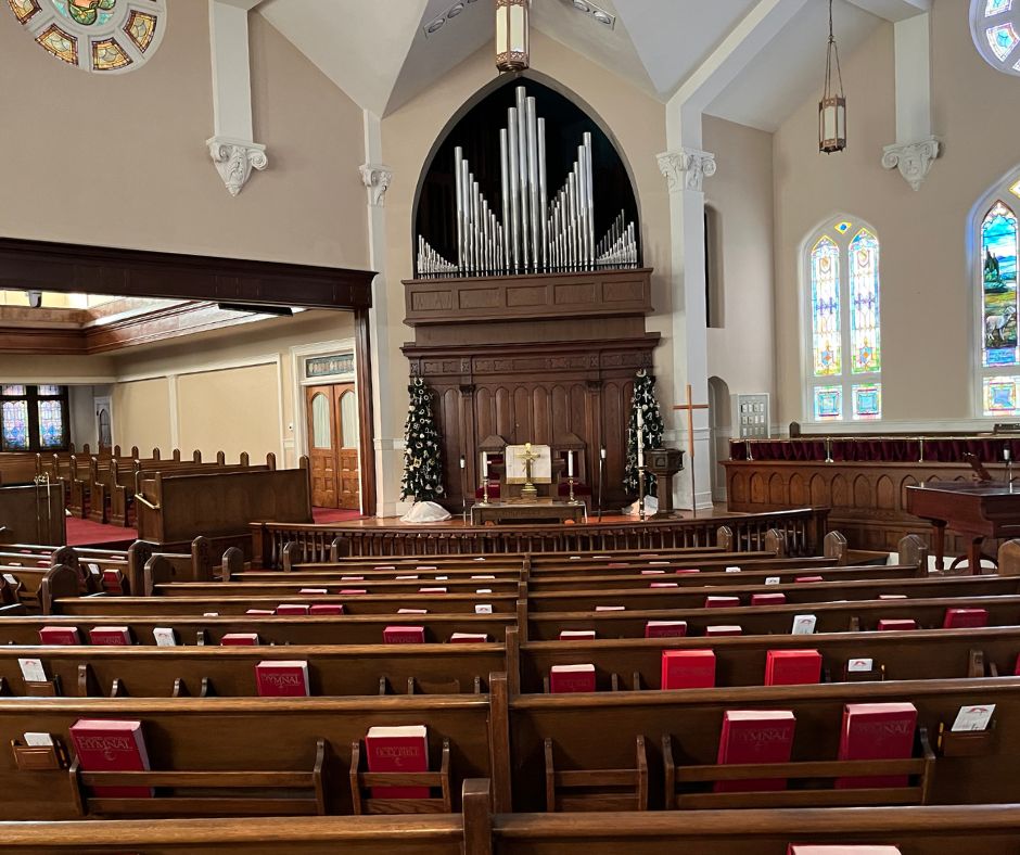 Traditional church sanctuary featuring dark wood pews with red hymnals, ornate pipe organ in arched alcove, altar with white flowers, and colorful stained glass windows